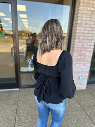 Woman wearing black long-sleeve blouse with tie-back detail and blue jeans, shown from behind.