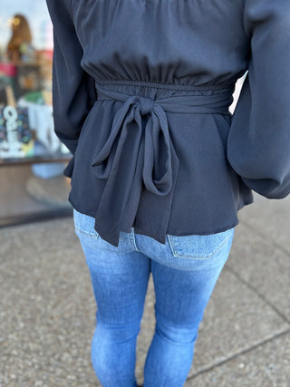 Woman wearing navy blue long-sleeve blouse with tie waist detail and blue jeans, shown from behind.