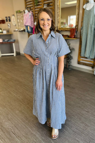 Woman wearing striped button-front maxi shirt dress with short sleeves in clothing boutique setting.