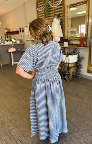 Woman wearing striped maxi dress with smocked waist, shown from behind in boutique setting.