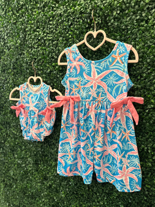 Two matching blue tropical print dresses with pink starfish pattern and coral ribbon belts on heart-shaped hangers.