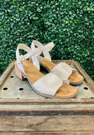 TOMS Eloise sandals with shimmer finish and ankle strap on wooden crate against greenery background