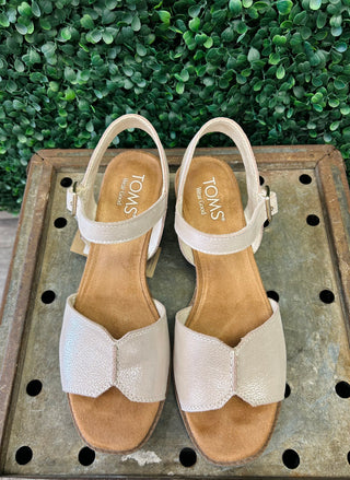 TOMS Eloise sandals in shimmer finish with ankle straps on wooden surface with greenery background