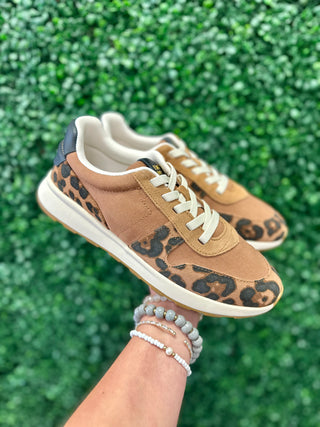 Hand holding brown leopard print TOMS sneakers with white laces against green foliage background