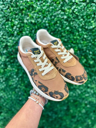 Hand holding pair of TOMS leopard print sneakers with brown tones and white laces against green background.