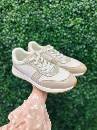 Hand holding TOMS retro sneakers with champagne shimmer finish against green foliage background.