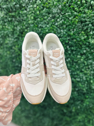 Hand holding pair of TOMS neutral-toned retro sneakers with white laces against green foliage background.