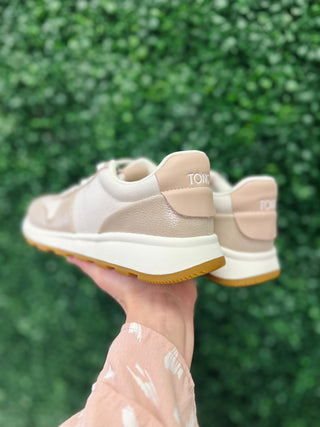 Hand holding beige TOMS sneakers with shimmer finish and gum sole against green foliage background.