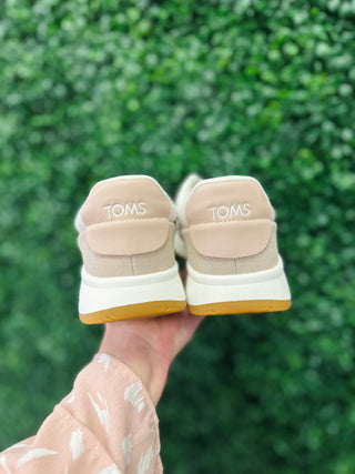 Hand holding pair of TOMS sneakers showing heel design against blurred green foliage background.