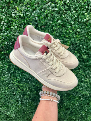 Hand holding TOMS retro sneakers with beige upper and pink heel accents against green foliage background