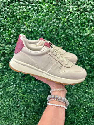 Hand holding beige TOMS sneakers with pink accents against green foliage background, wearing beaded bracelets.