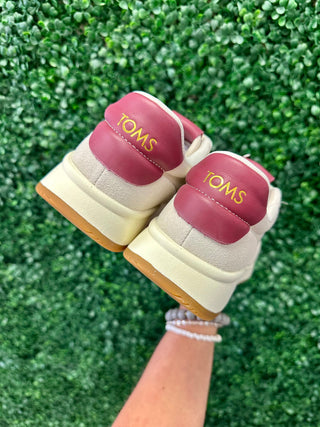 Hand holding pair of TOMS sneakers with pink leather accents against green foliage background