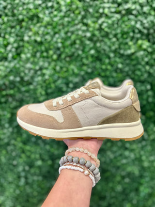 Hand holding beige suede sneaker with white sole against green foliage background, wearing beaded bracelets.