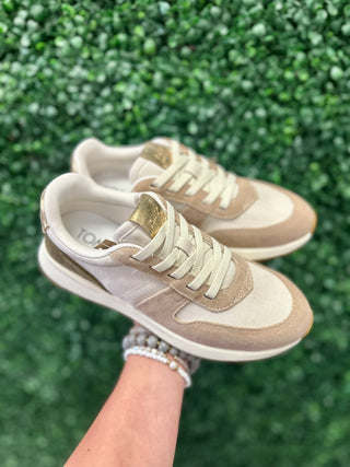 Hand holding pair of beige suede sneakers with white laces against green foliage background