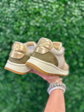 Hand holding TOMS retro suede sneakers with golden heel details against green foliage background.