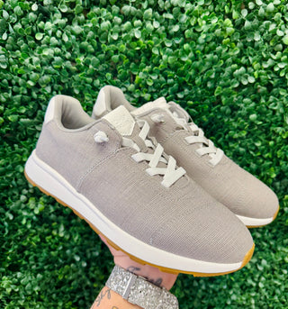 Hand holding gray TOMS sneakers with white laces and gum sole against green foliage background.