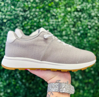 Hand holding gray canvas sneaker with white sole and gum rubber outsole against green foliage background