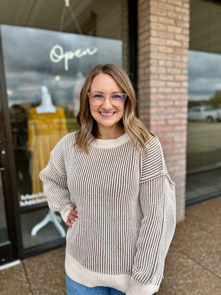 Woman wearing a ribbed knit crew neck sweater with two-tone striped pattern, standing outside boutique.