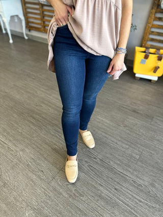 Woman wearing dark blue skinny jeans with button fly, pink top, and tan loafers in modern interior.