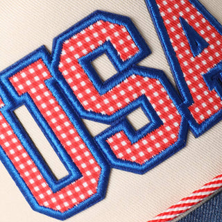 Close-up of embroidered USA letters in red gingham fabric with blue border on cream hat.