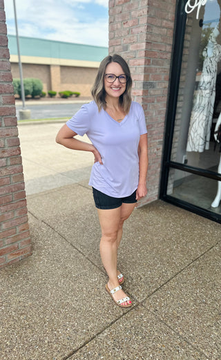 Woman wearing purple v-neck basic tee with dark shorts and sandals, standing outside boutique storefront.