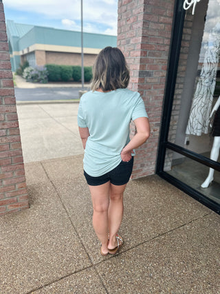 Woman wearing light blue v-neck basic tee and dark shorts, shown from behind on sidewalk.