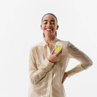 Smiling woman in wet beige shirt holding Touchland Vanilla Blossom hand sanitizer mist against white background.