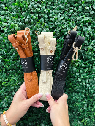 Hands holding three vegan leather bag straps with black packaging labels against green foliage background.
