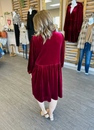 Woman wearing burgundy velvet babydoll dress with long sleeves in clothing boutique, shown from behind.