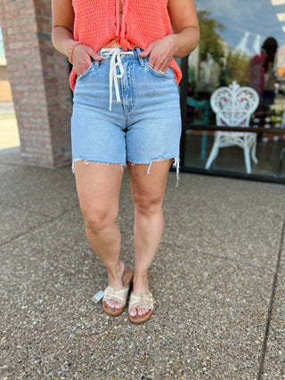 Woman wearing high-rise light wash denim shorts with drawstring waist and frayed hem, styled with coral top and sandals.