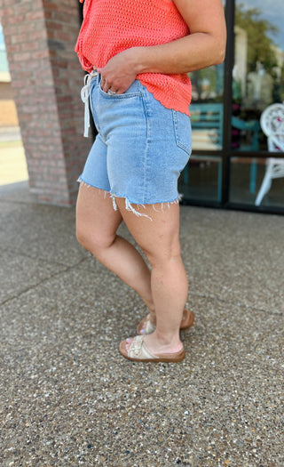 Woman wearing light wash denim drawstring shorts with frayed hem and coral knit top, standing outdoors.