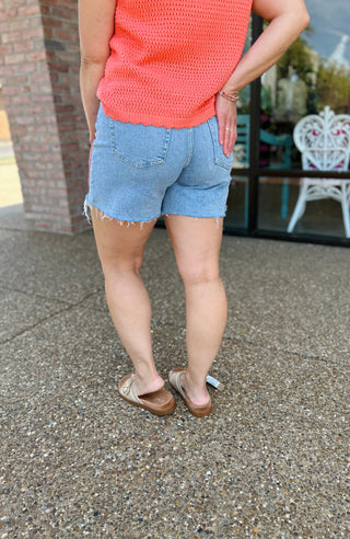 Woman wearing light wash denim drawstring shorts with frayed hem and coral knit top, back view.