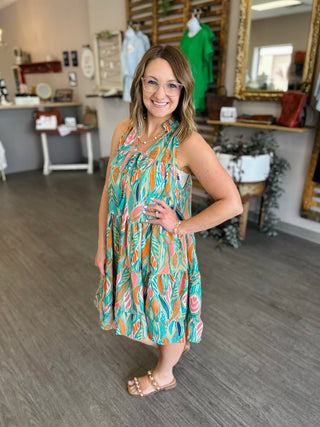 Woman wearing colorful tropical print sleeveless tiered dress with palm leaf pattern in retail boutique setting.