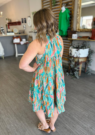 Woman wearing colorful tropical print sleeveless dress with flowing fabric, shown from behind in boutique setting.