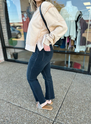 Woman wearing dark wash high-rise cropped flare jeans with frayed hem, paired with beige sweatshirt and sneakers.