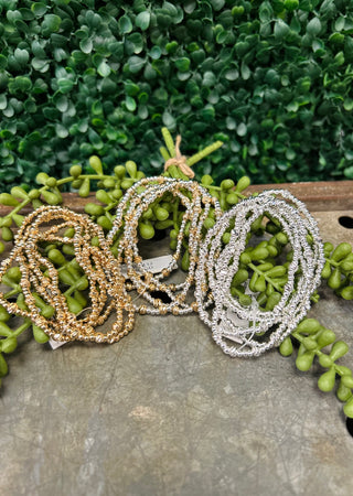 Stack of sparkly beaded bracelets in metallic tones displayed on wooden surface with green succulent plants.