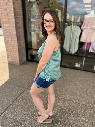 Woman wearing sage green sleeveless cami top with denim shorts, standing outside retail storefront.
