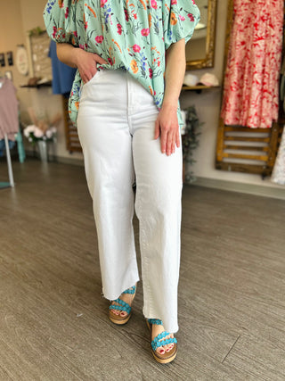Woman wearing white high-rise cropped wide leg jeans with floral top and blue sandals in clothing store.