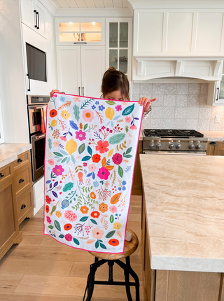 Woman displaying colorful wildflower patterned kitchen towel with vibrant floral design in modern kitchen setting.