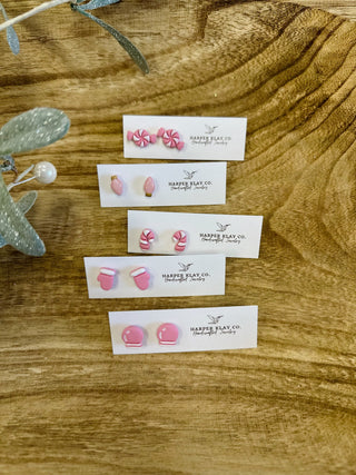 Five pairs of pink polymer clay stud earrings displayed on white cards on wooden surface.