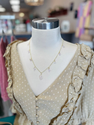 Mannequin wearing delicate gold chain necklace with dangling charms over beige polka dot ruffle top in boutique setting.
