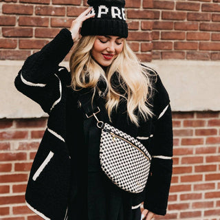 Woman wearing black striped sweater and beanie with checkered crossbody bum bag against brick wall