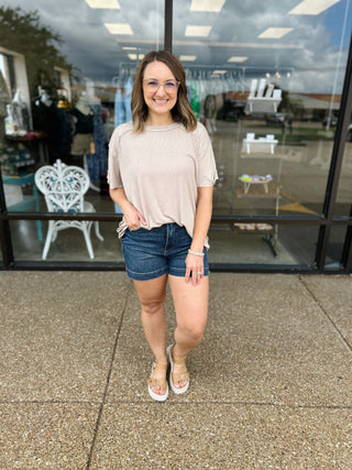 Woman wearing casual beige ribbed short sleeve top with denim shorts standing outside boutique storefront