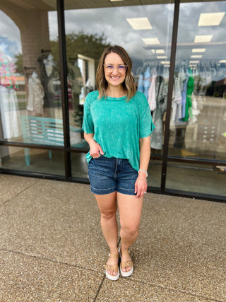Woman wearing casual green ribbed short sleeve top with denim shorts, standing outside clothing store