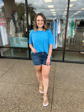 Woman wearing blue ribbed short sleeve top and denim shorts standing outside clothing store