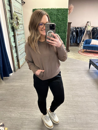 Woman wearing taupe sweatshirt and black cuffed joggers with zipper pockets taking mirror selfie in boutique.